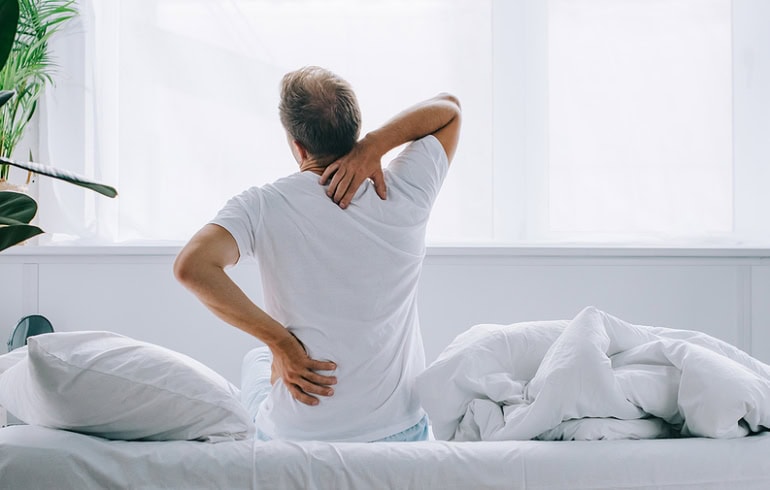 Man sitting on the bed facing away holding his shoulder and neck as if in pain.