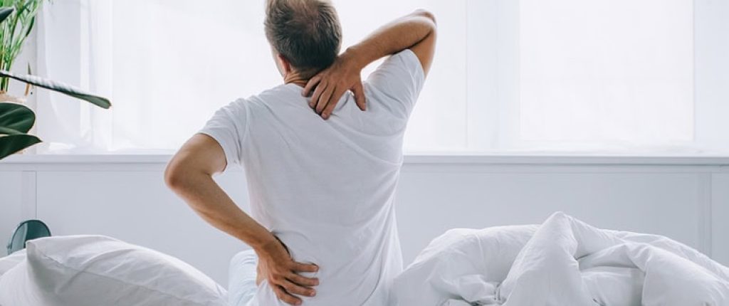 Man sitting on the bed facing away holding his shoulder and neck as if in pain.